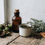 An old corked brown medicine bottle, an enamel cup, green black walnut hulls, a bundle of dried wormwood, clove buds and cinnamon on a farmhouse windowsill