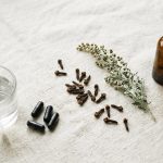 A glass of water with activated charcoal capsules set apart from an amber tincture bottle, with clove buds and wormwood between them