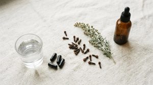 A glass of water with activated charcoal capsules set apart from an amber tincture bottle, with clove buds and wormwood between them